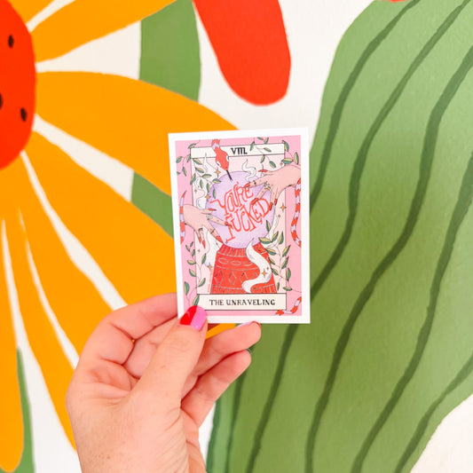 A hand with tattoos holds a tarot card titled 'The Unskilling' in front of a colorful mural featuring a large flower and green leaf shapes.
