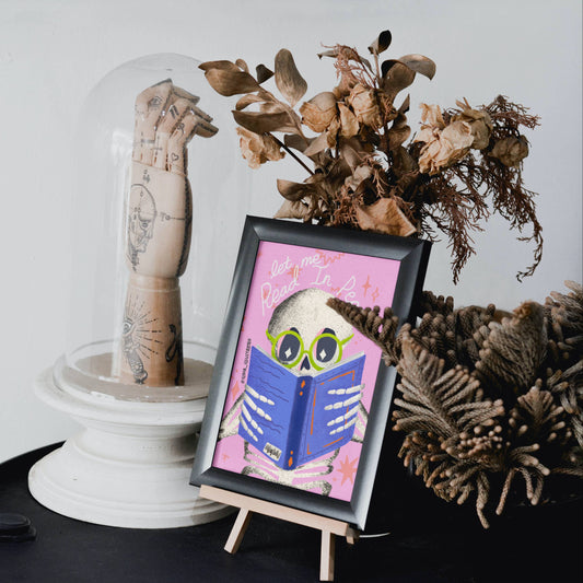A framed art print featuring a cartoon skull wearing green goggles and reading a blue book on a pink background, displayed on a small wooden easel. The scene is styled with dried botanicals and a glass dome covering a tattooed hand sculpture, set against a clean white background.