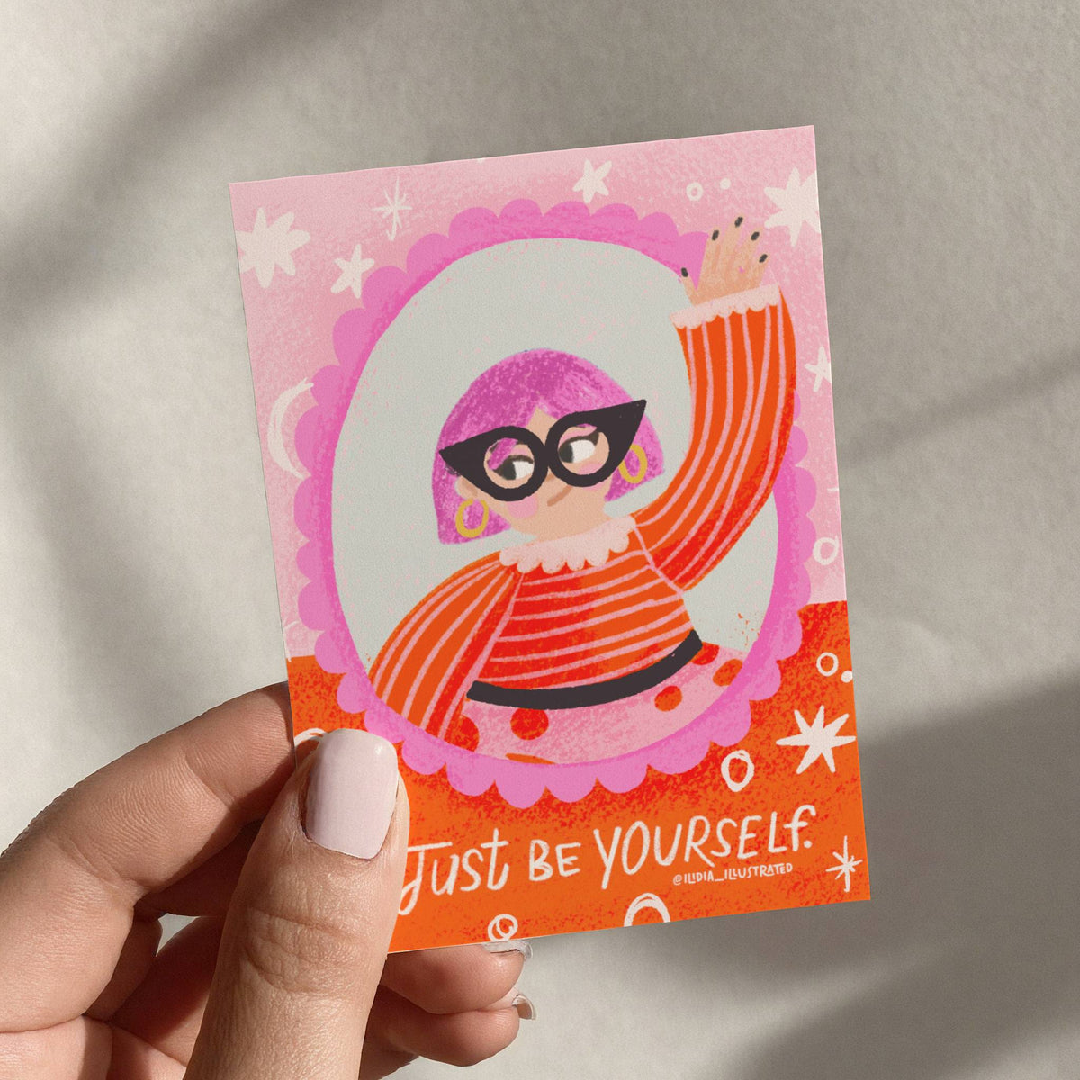 Hand holding a pink and orange card with a stylized illustration and 'Just Be Yourself' text.