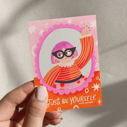 Hand holding a pink and orange card with a stylized illustration and 'Just Be Yourself' text.