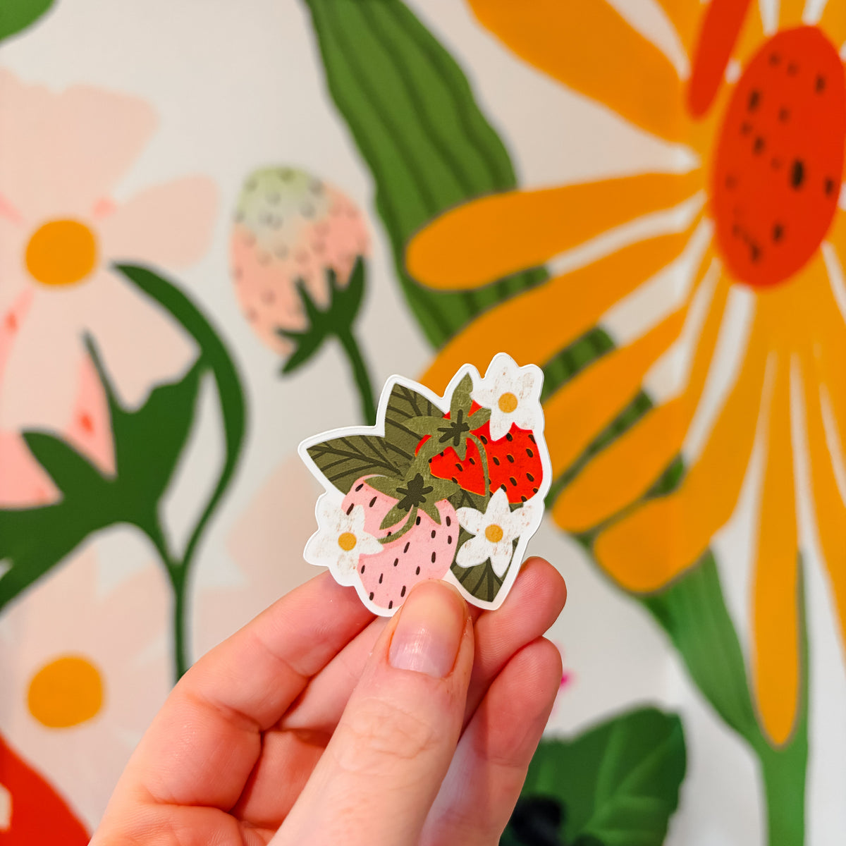 Hand holding a small sticker with strawberry design against a floral background