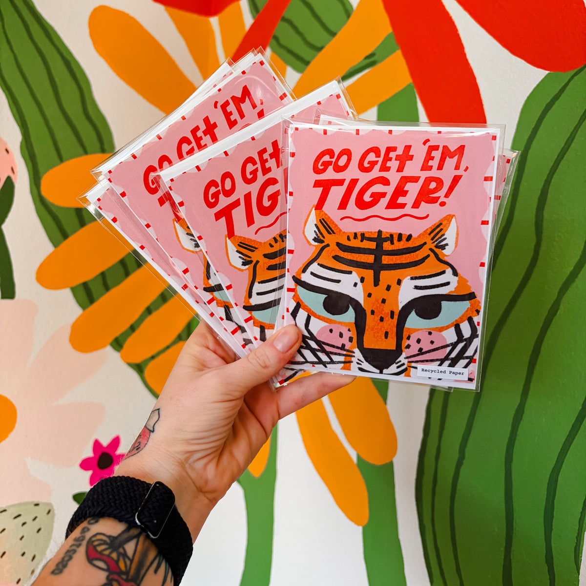 Hand holding two 'Go Get Em Tiger!' cards against a colorful floral and cactus background.