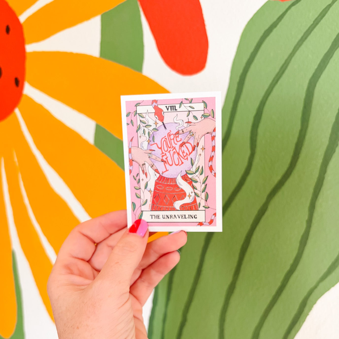 A hand with tattoos holds a tarot card titled 'The Unskilling' in front of a colorful mural featuring a large flower and green leaf shapes.