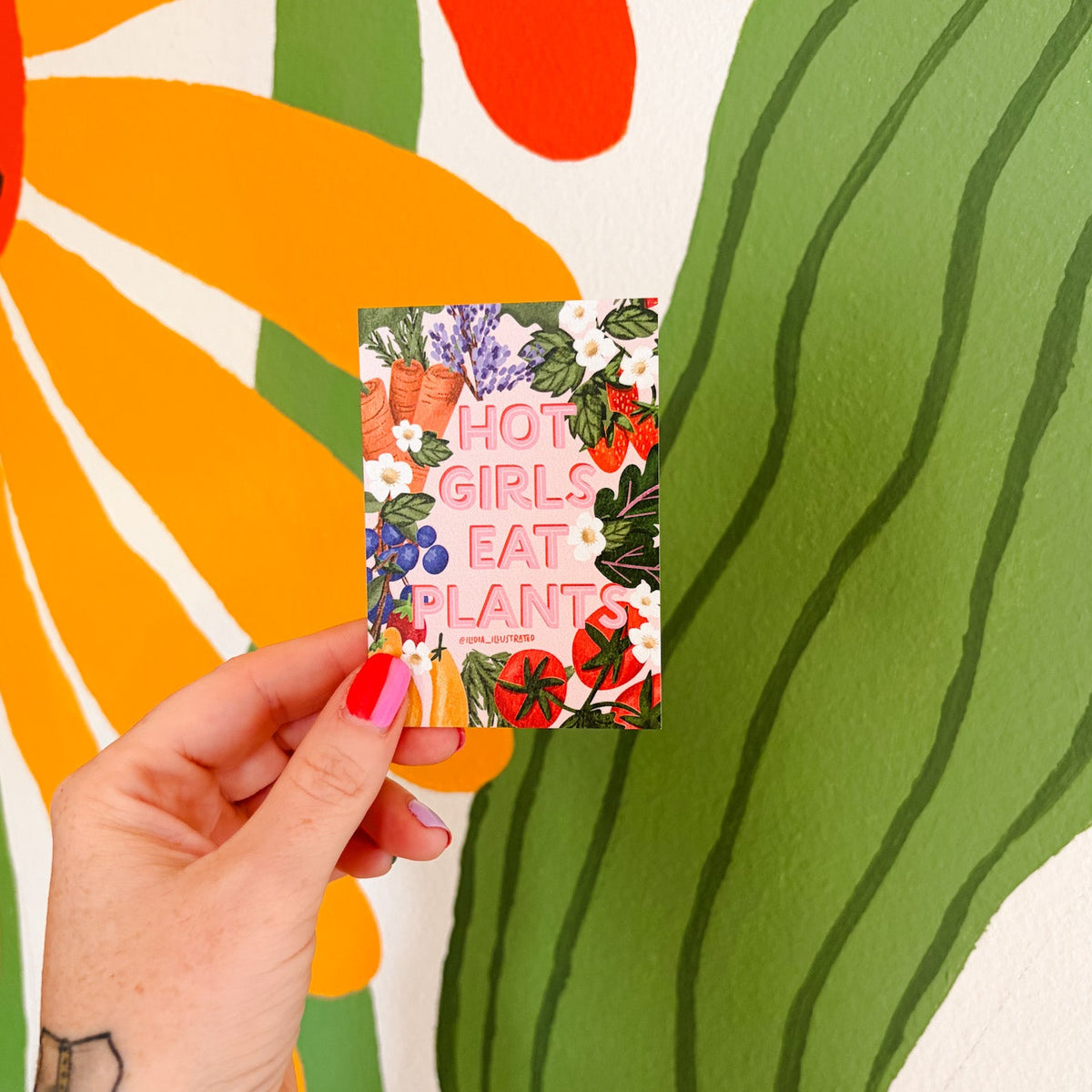 A hand with red nail polish and a strawberry tattoo holds a small square card that reads 'HOT GIRLS EAT PLANTS' in pink letters, set against a large colorful mural of a sunflower and a green cactus.