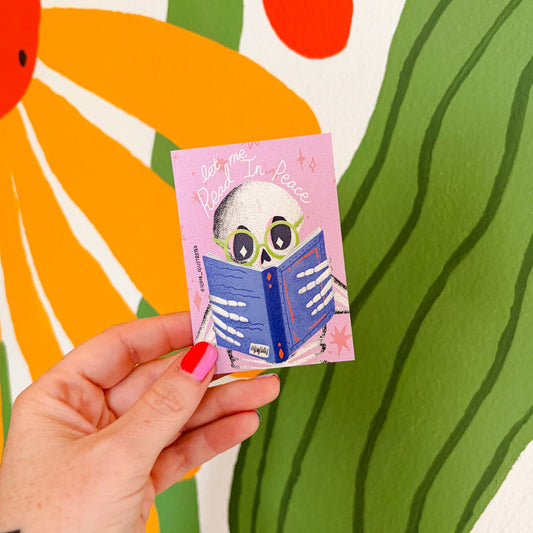 A hand with red nail polish holds a small pink card featuring a cartoon skull reading a blue book. The background is a large colorful mural with a yellow sun and green leaf shapes.