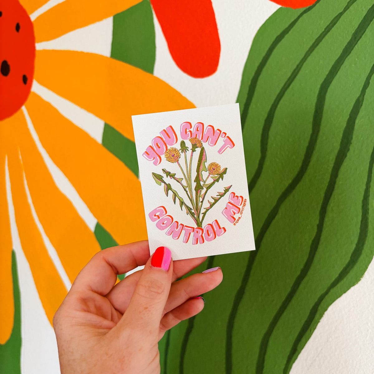 A hand with bright pink nail polish holds a small white art print featuring pink text that reads 'YOU CAN'T CONTROL ME' around a green and orange thistle illustration. The background is a large, colorful mural with abstract floral shapes in orange, green, red, and white.