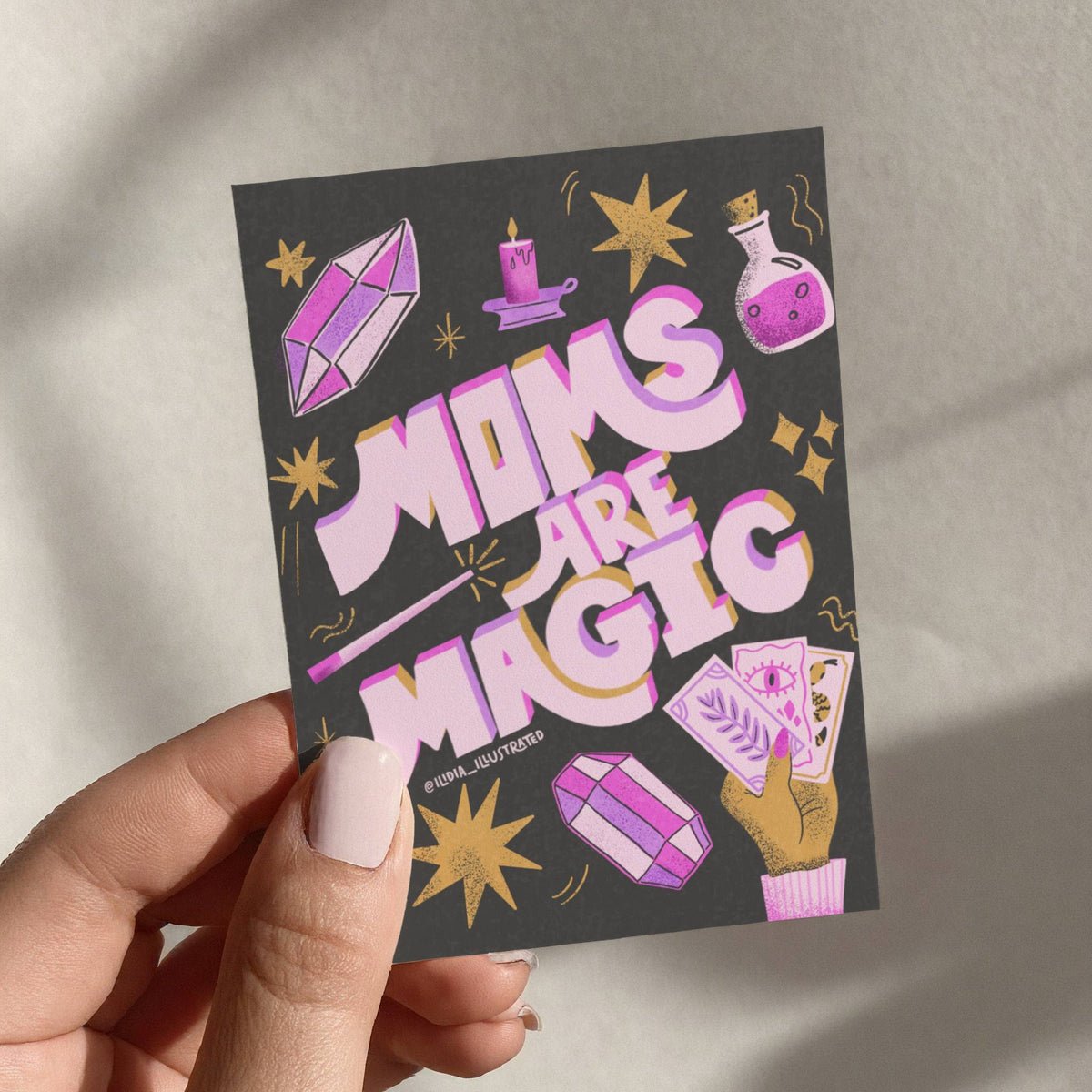 Hand holding a card with 'Moms are Magic' text and magickal illustrations.