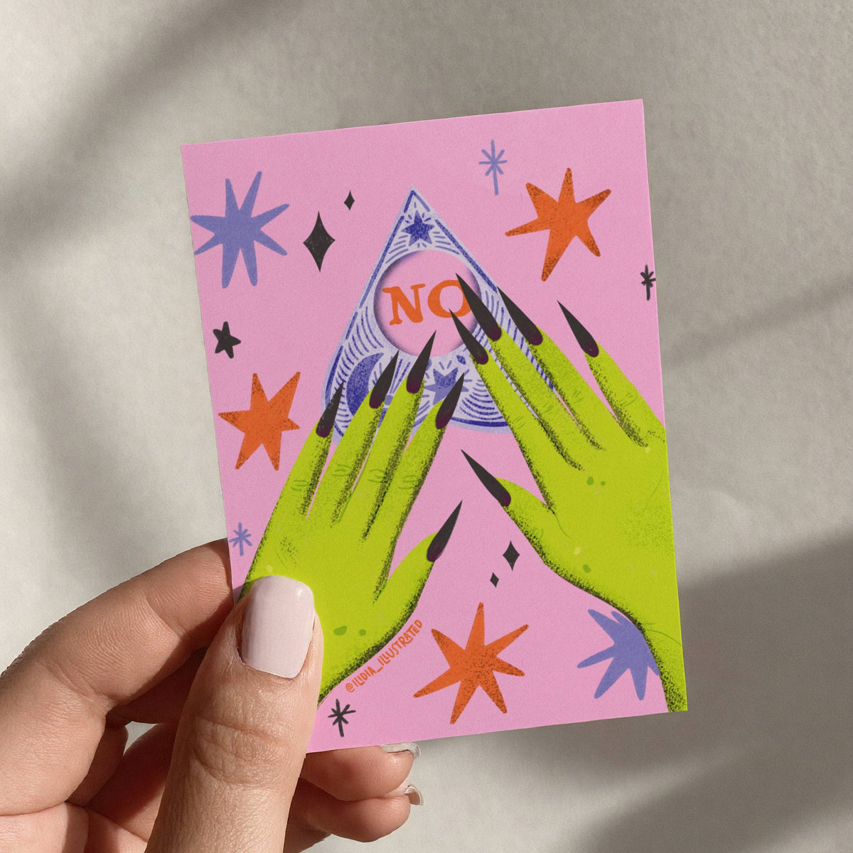 A hand holding a pink art print or card featuring a stylized green hand with long black nails in front of a blue mountain with the word 'NO' in orange, surrounded by colorful star shapes.