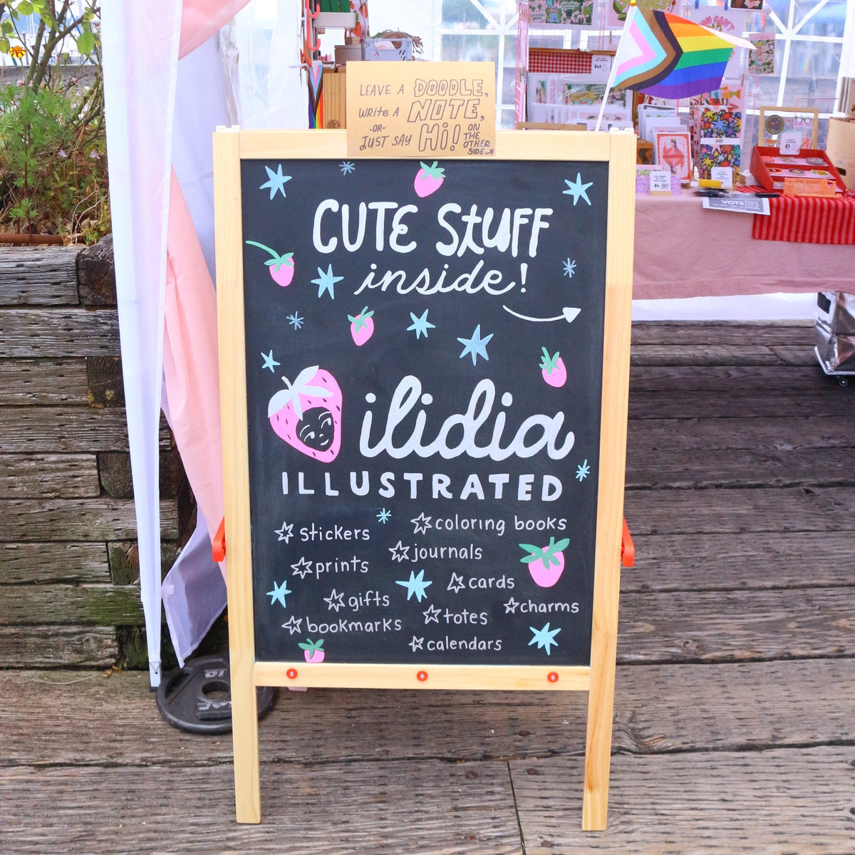 A wooden A-frame chalkboard sign on a wooden deck at an outdoor market. The sign reads 'CUTE SKUFF inside! ilidia ILLUSTRATED' with pink and blue doodles, and a small sign behind it says 'I LOVE A BARBIE BOOK CLUB'.