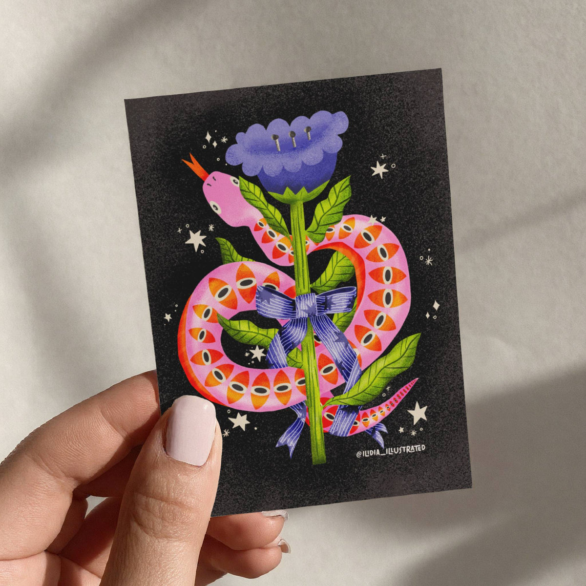 Hand holding a postcard with a colorful illustration of a snake holding a flower on a black background.