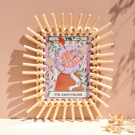 A square art print featuring the 'The Unraveling' tarot card illustration, framed by a circular arrangement of small wooden clothespins on a beige surface against a peach-pink wall.