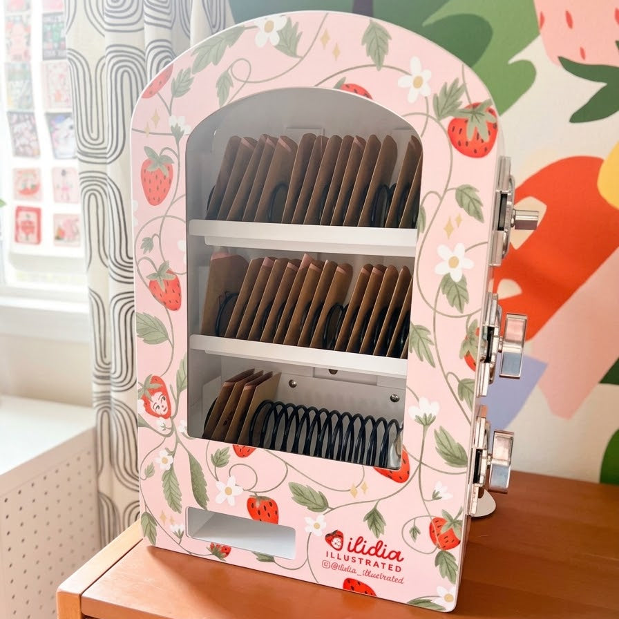 A strawberry-themed tea dispenser by ilidia on a wooden table, with a colorful illustrated wall and window in the background.