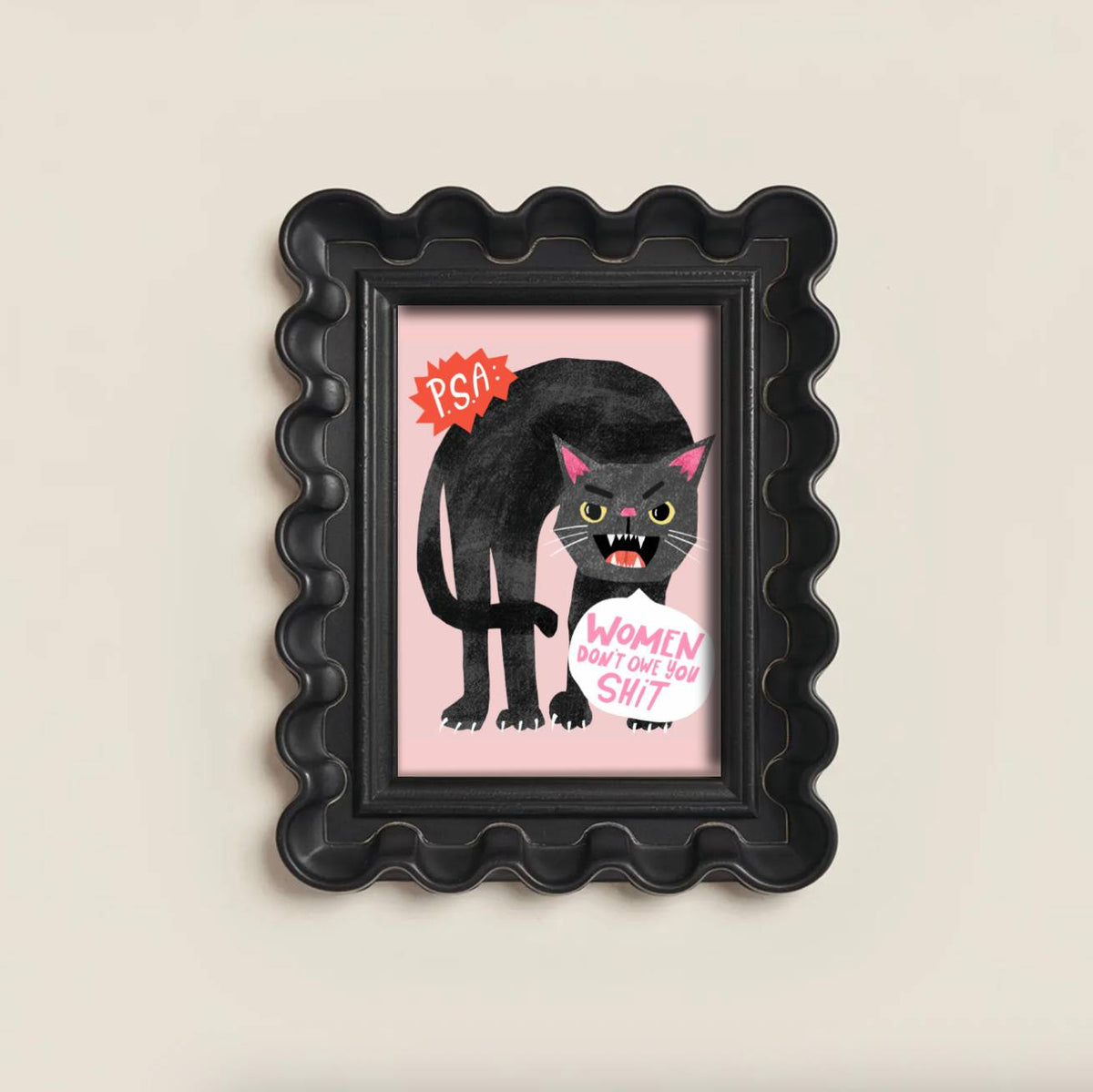 A black scalloped picture frame containing a cartoon illustration of a black cat with pink ears holding a white sign that reads 'WOMEN DON'T LOVE YOU SHIT' on a light pink background, shown against a warm off-white wall.