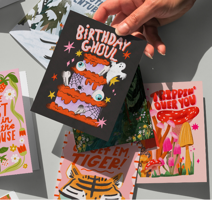 A hand holds a black birthday card with a colorful cake illustration, next to several other illustrated cards. The right side shows a pink background with the text 'little pieces of hand-drawn art for your everyday life' and an orange 'shop now' button, plus a small strawberry drawing above.