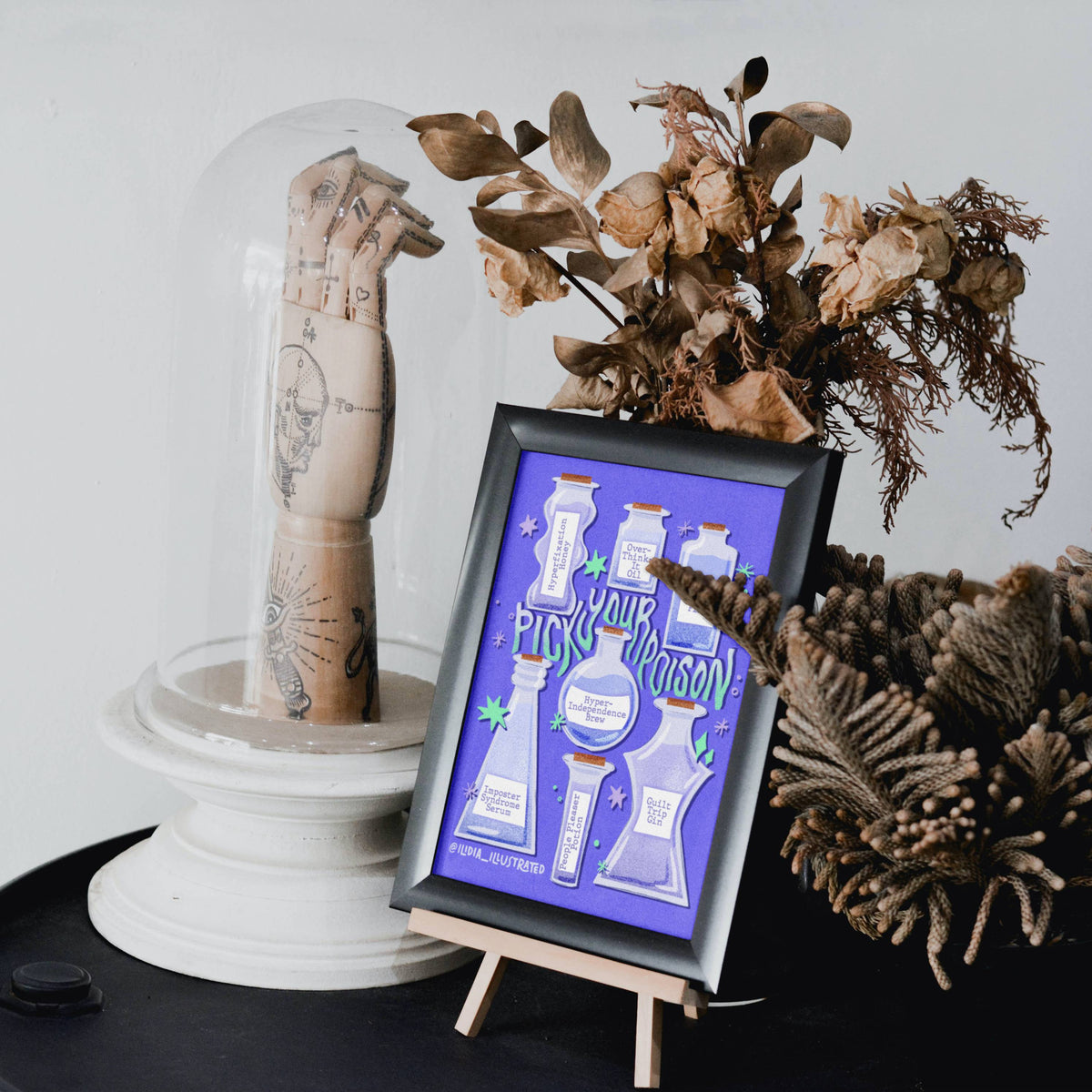 A framed art print featuring white illustrations of laboratory glassware on a purple background, styled on a small wooden easel next to a glass dome with a tattooed arm sculpture and dried botanicals.