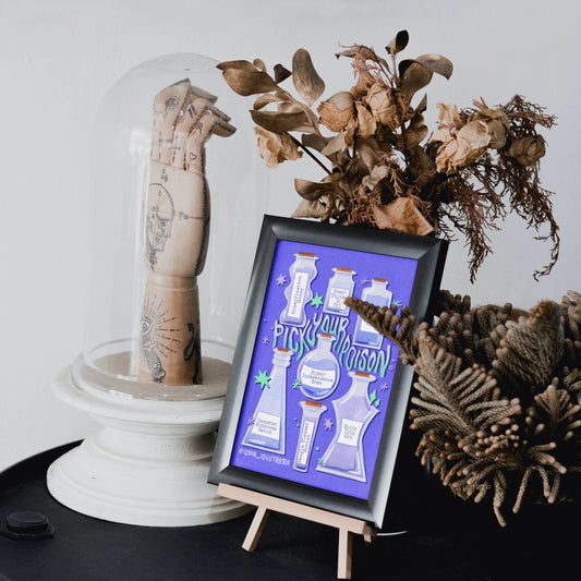 A framed art print featuring white illustrations of laboratory glassware on a purple background, styled on a small wooden easel next to a glass dome with a tattooed arm sculpture and dried botanicals.