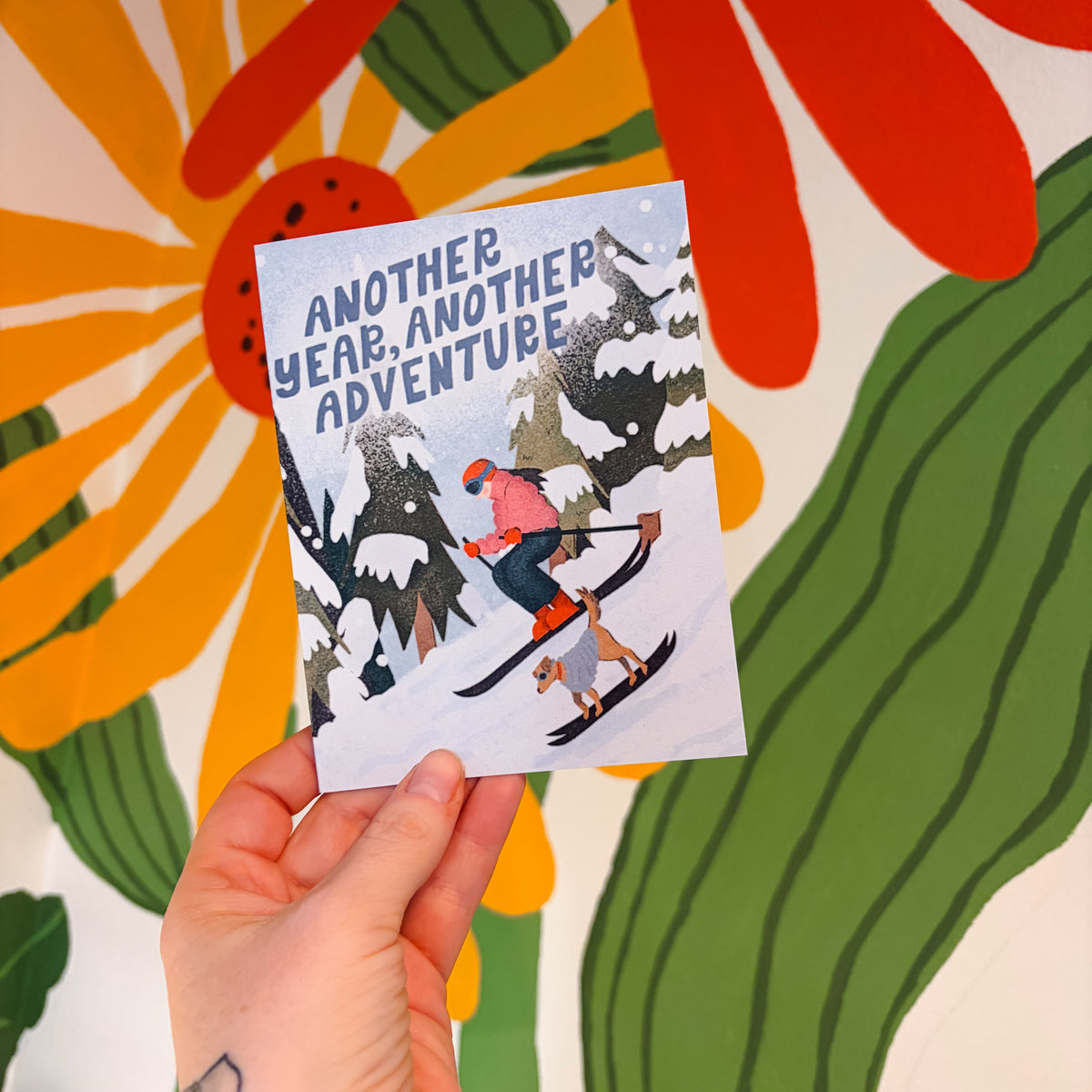 Hand holding a card with skiers and 'Another Year, Another Adventure' text against a colorful floral background.