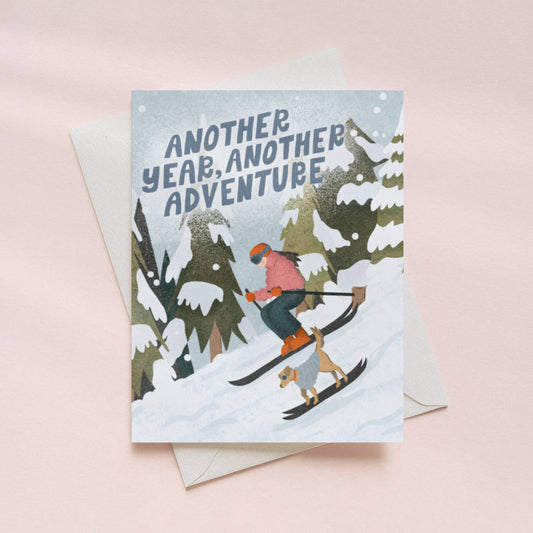 an illustration of a woman and her best friend dog skiing down the alpine slopes with text 'another year, another adventure' birthday card.