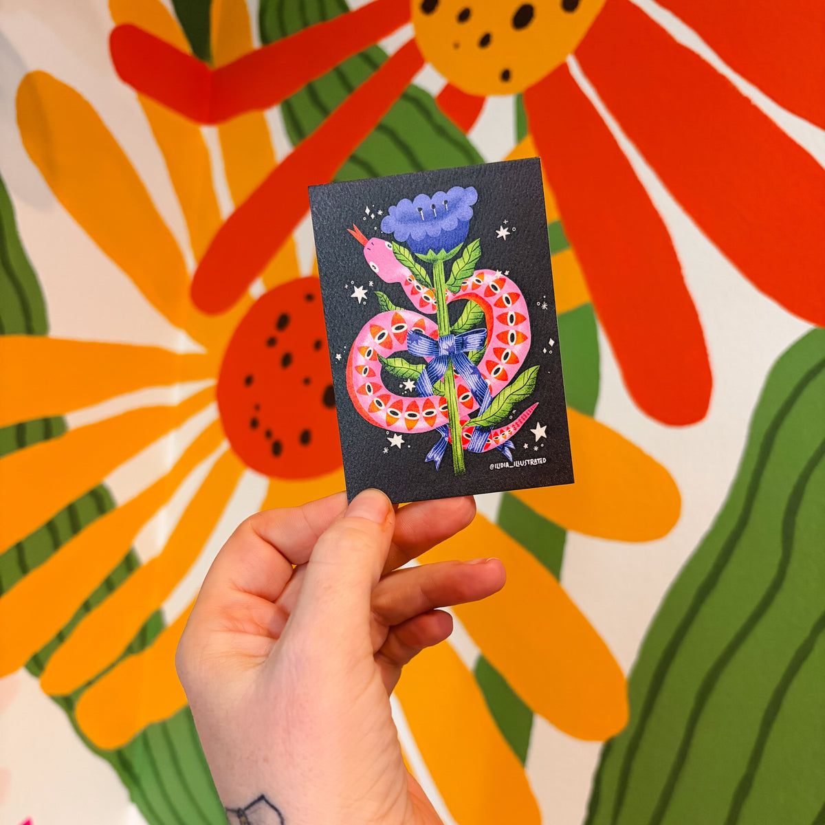 Hand holding a card with a colorful design of a snake and flowers against a floral background