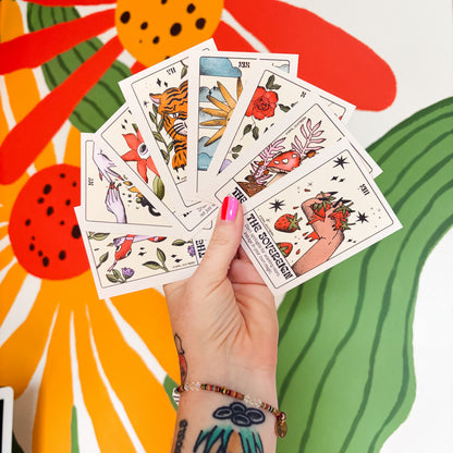 A hand with colorful tattoos and pink nail polish holds a fan of illustrated tarot cards, including The Fool and The Hanged Man, against a vibrant, stylized floral background.